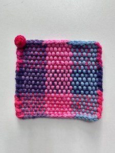 Close-up of mug rug in colors of reds, pink,s blues, and purples. Plastic rosebud button at upper left-hand corner.