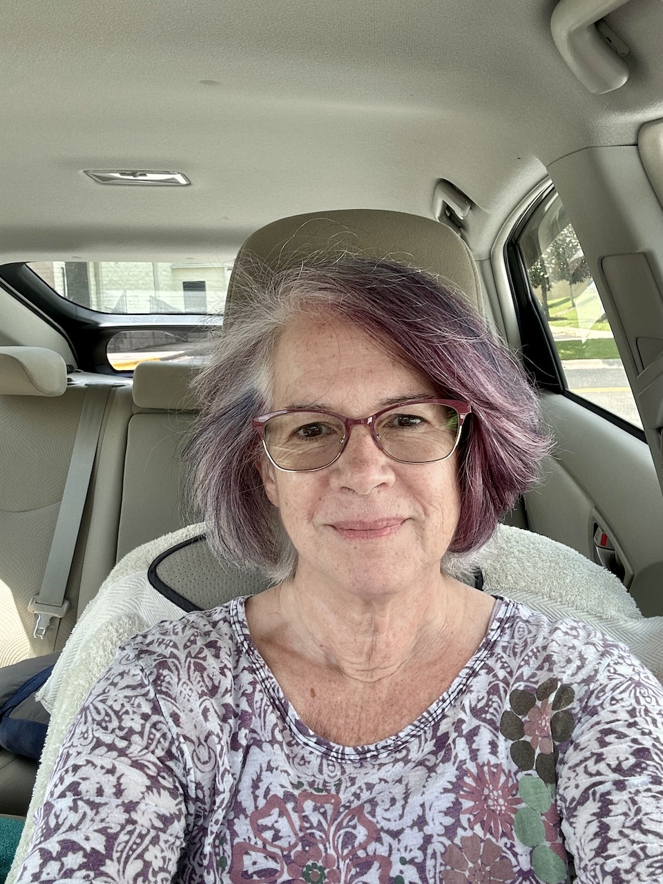 Woman in her sixties, wearing purple-framed glasses and sporting a purple and slightly blue haircut (almost shoulder length). Sitting in her car and wearing a floral shirt that nearly matches her hair.