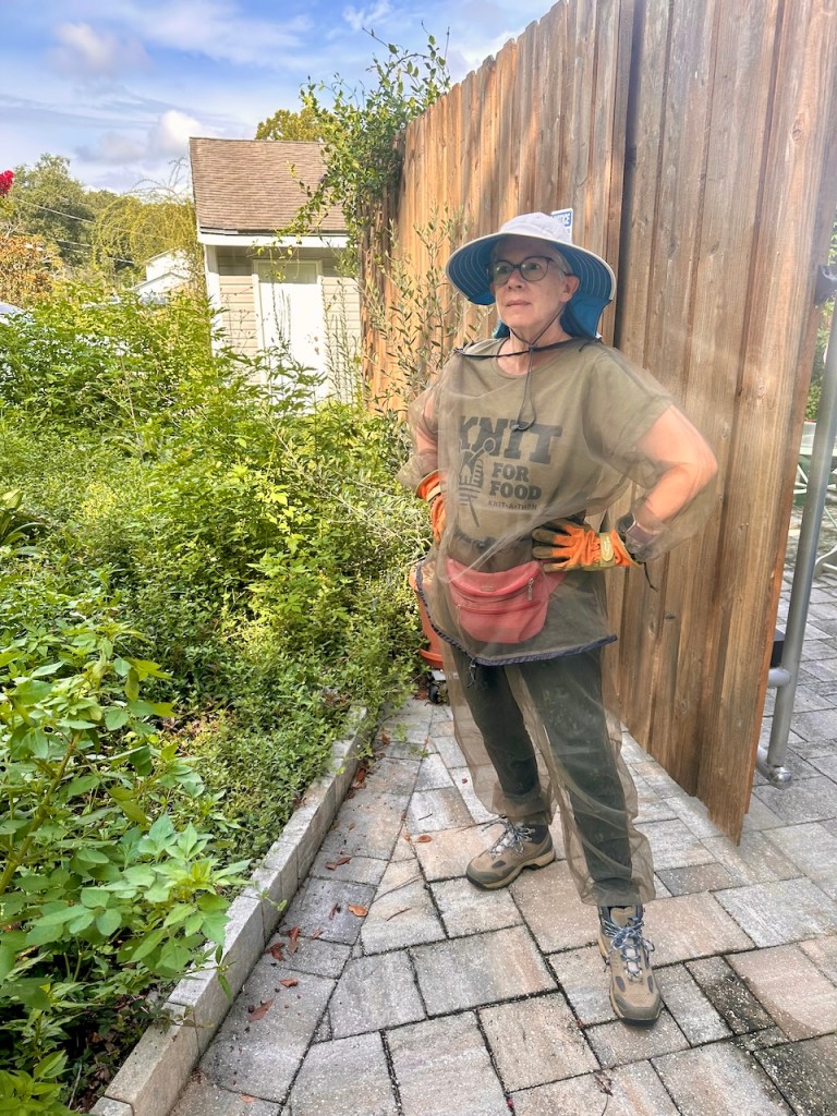 Adult woman, standing in front of a tall fence, dressed in long pants, a mesh pullover and wide-brim hat. 