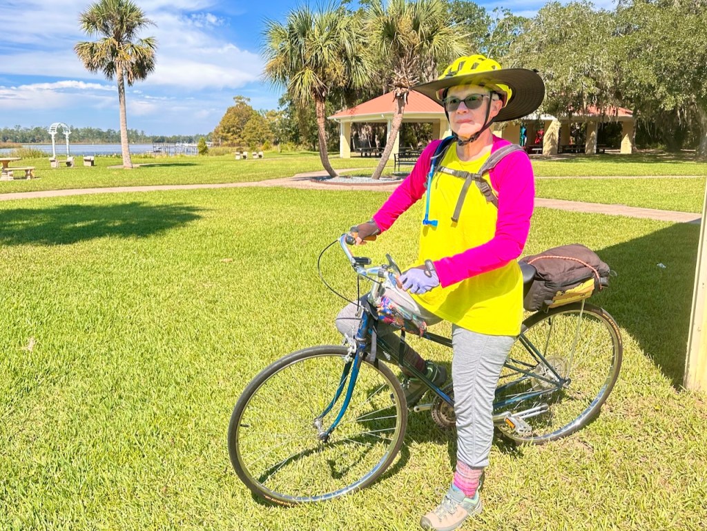 Adult woman on a bicycle wearing gray leggings, bright yellow sleeveless top,  a bright pink bolero, and a bright yellow helmet with wide brim.